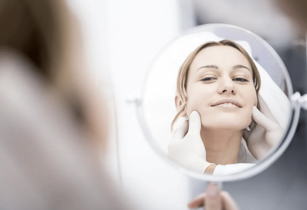 A patient studies her chin and jawline in a hand mirror while a gloved surgeon supports her jaw on both sides